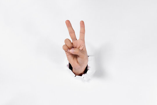 Man's hand showing a peace sign through a torn white paper hole, victory and encouragement concept.