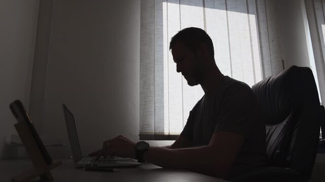 Side view silhouette of a young businessman working on a laptop at a office desk by the window