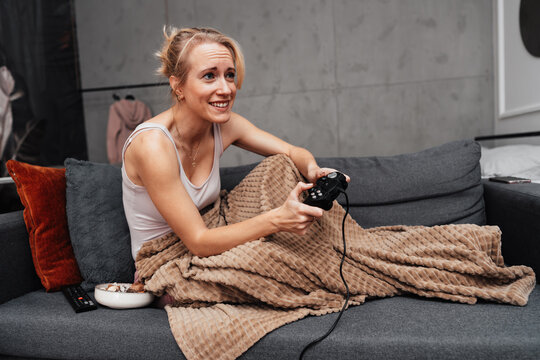 A cheerful female gamer relaxes under a warm blanket while holding a gamepad. She enjoys a fun gaming session on a comfortable grey sofa with snacks nearby.