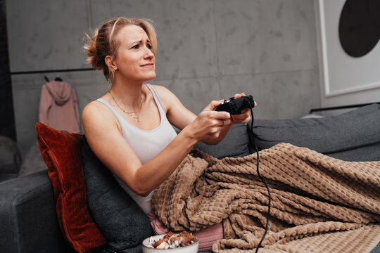 A stressed woman reacts to losing a match while playing video games in her cozy living room. She holds a gamepad and touches her head in frustration.