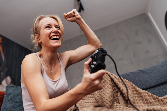 A cheerful female gamer laughs and raises her fist in triumph while holding a controller on her living room sofa.