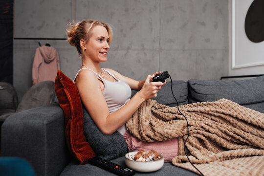 A cheerful female gamer relaxes under a warm blanket while holding a gamepad. She enjoys a fun gaming session on a comfortable grey sofa with snacks nearby.