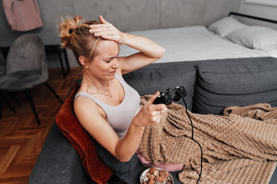 A stressed woman reacts to losing a match while playing video games in her cozy living room. She holds a gamepad and touches her head in frustration.