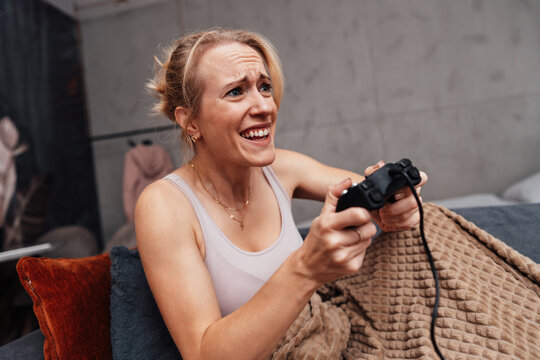 An intense female gamer reacts with a passionate grimace while holding a black controller under a cozy blanket on the sofa.