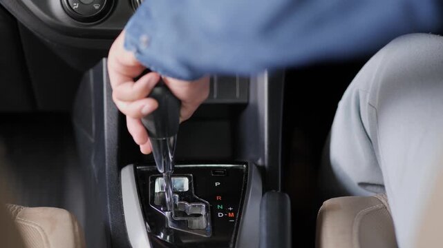 Close-up shot of a driver's hand shifting the automatic gear stick inside a car, with the gear panel showing P, R, N, D, and M positions