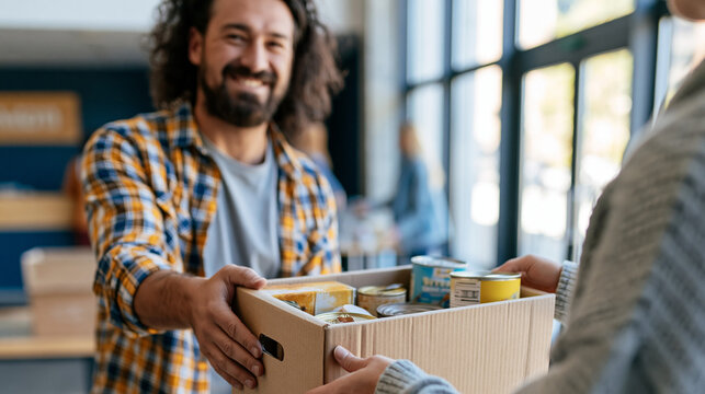 Smiling man handing a cardboard box of donated items to another person indoors, with books, supplies and community shelves in the background.
