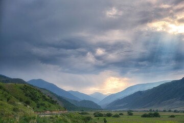 Fototapeta premium Stormy mountain valley landscape with winding road and sun rays breaking through dramatic clouds. Ideal for travel advertising, background, and nature editorial usage with copy space.