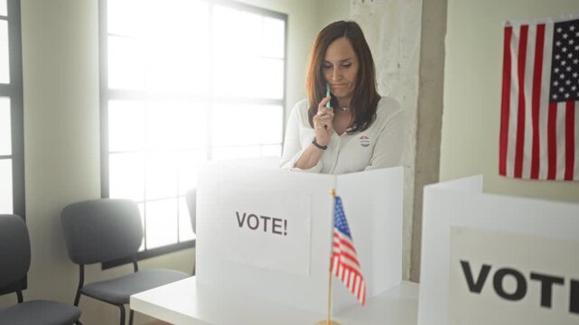 Woman at voting booth holds pen to chin and studies ballot in polling building; civic duty thoughtful.