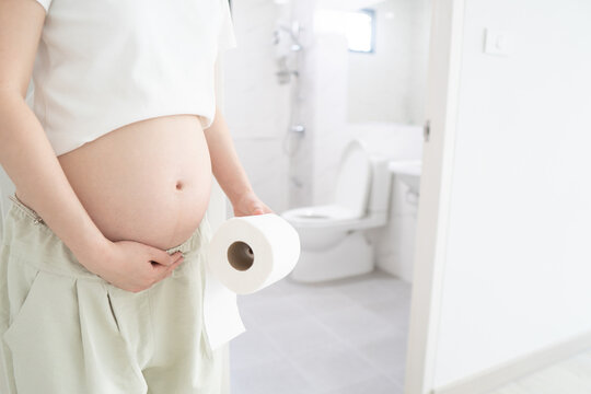 Constipation during pregnancy concept. Pregnant woman suffering form abdominal pain and holding​ toilet tissue​ paper roll in front of the bathroom at home.