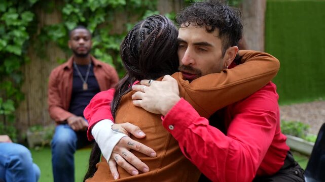 Two people are hugging each other tightly, providing mutual support and comfort during an emotional group therapy session, while other members of the group watch and listen in the background