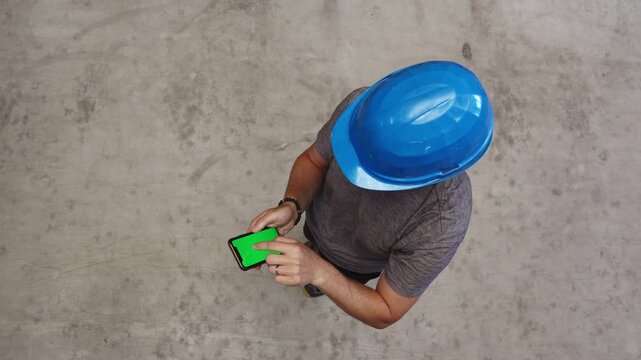 Top down view of a worker wearing a blue hard hat using a smartphone with a green screen on a concrete background