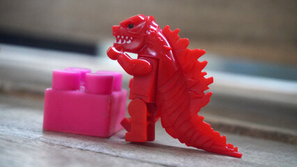 Red toy dinosaur figure standing beside a pink building block on a wooden surface. © anungcamui