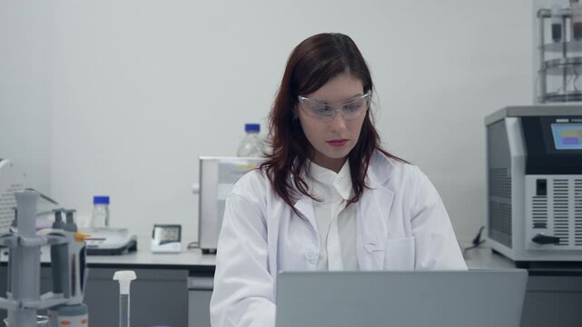 Female scientist wearing lab coat and safety glasses working on laptop in modern laboratory research facility