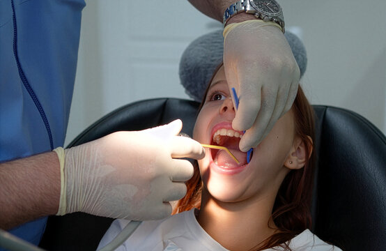 Close up of a male dentist in medical gloves using a dental mirror and air water syringe tool to check and clean a young girl's open mouth during a checkup in a dental chair.
