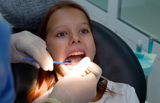 Close up high angle shot of a professional dentist in medical gloves using a dental mirror and probe tool to examine teeth of a young girl patient lying in a black dental chair at a clinic.