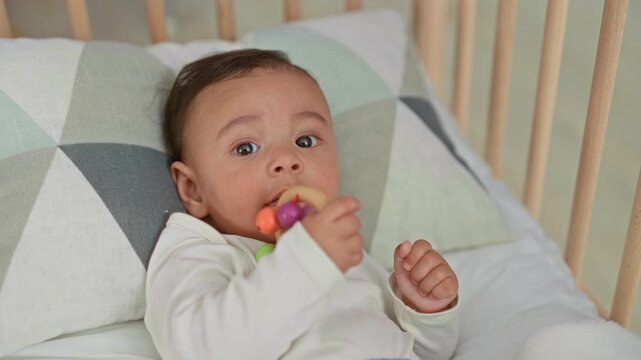 Baby boy holds and mouths a colorful teether while lying in a crib inside a building; curiosity comfort.