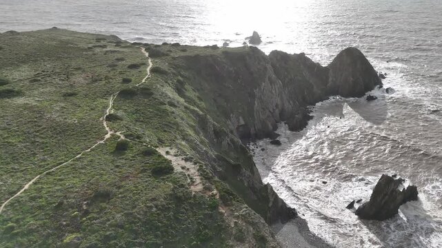 Aerial drone push in shot of the rugged coastal landscape of the Fisherman's Trail, Rota Vicentina, in Portugal. Lush green cliffs meet the Atlantic Ocean with crashing waves at sunset.