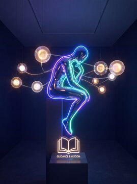 Neon art installation of a human figure sitting with legs crossed inside a dark room, illuminated by glowing orbs and an open book labeled "Guidance & Wisdom"