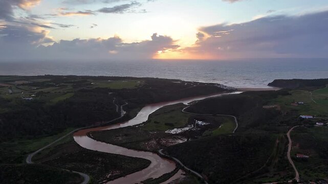 The video shows a push-in drone shot of a stunning river estuary meeting the Atlantic Ocean at sunset in Portugal, near Odeceixe. Lush green hills and a sandy beach complete the picturesque scene.