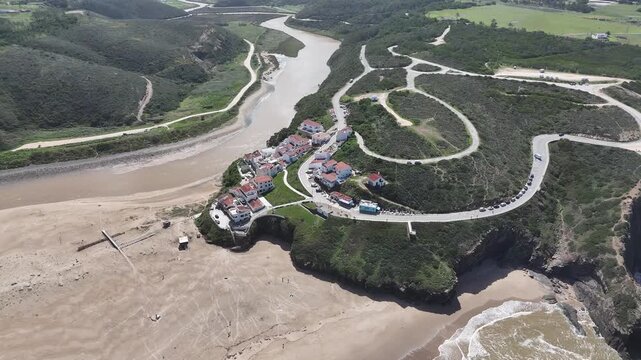 Aerial drone pan right and tracking shot over Praia de Odeceixe in Portugal. The river estuary meets the Atlantic coast, featuring sandy beaches, lush hills, and winding roads.