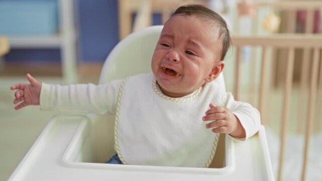 Baby boy crying and spitting spoonful of puree into mouth with messy bib while seated in highchair in building; feeding frustration.