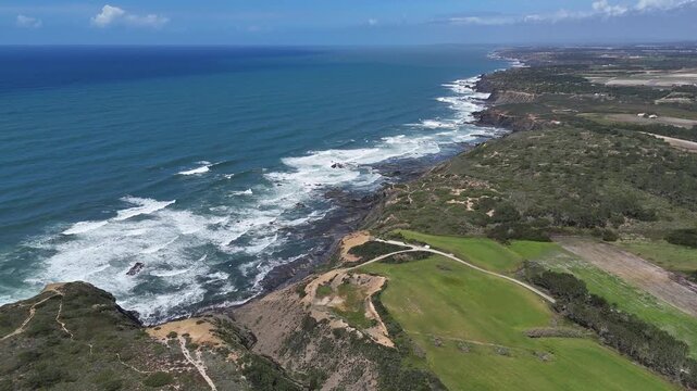 Aerial drone shot panning left along Portugal's beautiful rugged coastline near Rogil, featuring dramatic cliffs, crashing waves, and green fields. Part of the iconic Fisherman's Trail.