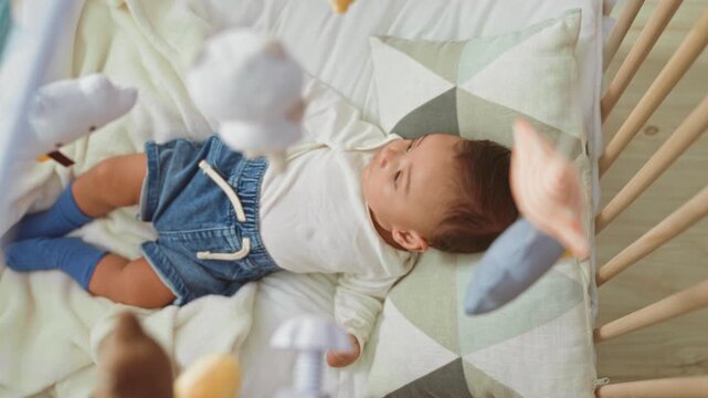 Baby boy lying on back in wooden crib bed, wearing denim shorts and blue socks, hand reaching toward hanging plush mobile above, pillow at head, floor visible in building; gentle curiosity.