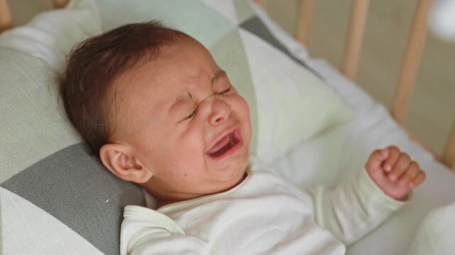 Baby boy crying in a wooden cradle, tearful face and raised hand visible in a nursery with patterned pillow; distress need comfort.