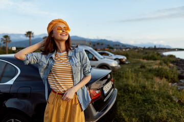 Fototapeta premium A smiling woman in a striped shirt, denim jacket, and mustard skirt poses beside a parked car in a sunny coastal landscape, embodying carefree, stylish outdoor living and spontaneous travel.