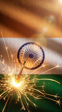 An Indian flag with fireworks sparkler burning in the foreground, casting warm golden light over a soft focus tricolor backdrop