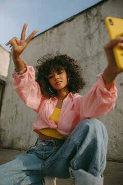 An attractive young woman in jeans and a pink shirt, doing a pose from breakdancing while taking a selfie with a yellow phone.