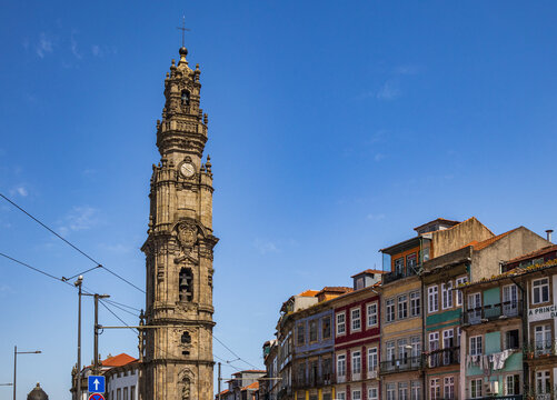 Vista desde un &aacute;ngulo bajo de la emblem&aacute;tica Torre de los Cl&eacute;rigos de estilo barroco, flanqueada por edificios residenciales con coloridas fachadas t&iacute;picas bajo un cielo azul en Oporto, Portugal.
