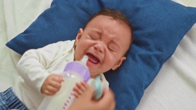 Baby boy holding milk bottle and feeding while crying on bed in building; upset soothing comfort care.