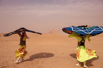Fototapeta premium Dual tanoura performance dancers in neon yellow robes spinning enormous painted discs on Giza desert plateau with iconic pyramids centered between