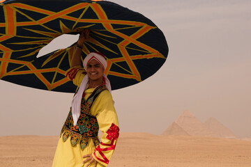 Fototapeta premium Smiling young tanoura dancer in ornate yellow Egyptian folk costume spinning colorful star patterned disc against hazy Giza pyramids desert landscape