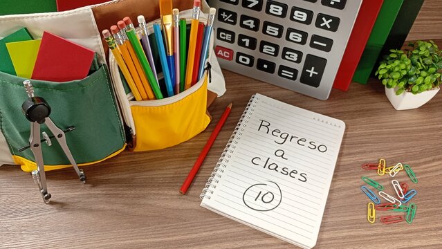Back to school concept featuring colorful school supplies arranged on a wooden desk, including notebooks, pencils, calculator, backpack and a spiral notebook displaying the message "Regreso a clases".