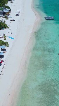 Aerial beach swing and swimmers in turquoise shallows, floating platform anchors playful activity, families wading near coral flats, white crescent sand and sunny coastal mood, Maldives,vertical video