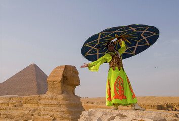 Fototapeta premium Sphinx performer in vivid neon robe raises large teal decorative disc overhead beside Great Sphinx at Giza plateau desert Egypt cultural tourism