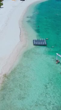 Aerial beach swing and swimmers in turquoise shallows, floating platform anchors playful activity, families wading near coral flats, white crescent sand and sunny coastal mood, Maldives,vertical video