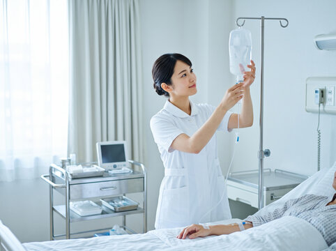 病室でベッドの患者に付き添い点滴を調整し確認する看護師の女性
