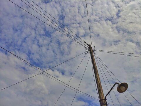 Wooden utility pole with messy electric wires and street lamp under cloudy blue sky