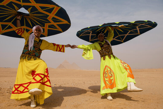 Two Egyptian tanoura folk dancers in vivid embroidered robes hold hands spinning colorful skirts overhead at the Giza Pyramids in Egypt ideal for