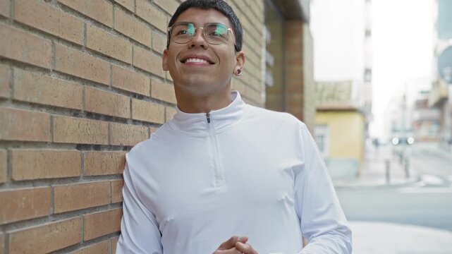 Man wearing glasses leans against a brick building, hands clasped and smiling in a casual white pullover on a city street; confidence.