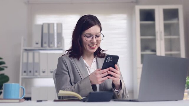 Businesswoman laughing and scrolling on her smartphone while showing different emotions sitting at her office desk