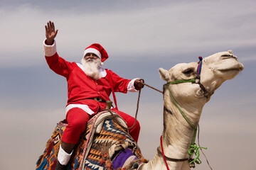 Fototapeta premium Santa waving enthusiastically from atop a white camel with vivid woven saddle blanket under pale desert sky ideal for global Christmas and