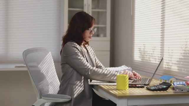Businesswoman working on laptop at sunny office desk drinking coffee while typing on keyboard