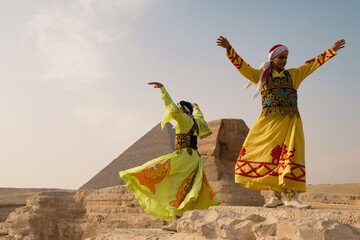 Obraz premium Two exuberant folk dancers wearing vivid embroidered yellow robes leap and swirl with arms raised beside the Great Sphinx and Great Pyramid of Giza