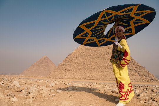 Folk costume clad boy smiling confidently while holding a bold geometric umbrella against the iconic Giza Pyramids in Egypt ideal for cultural tourism