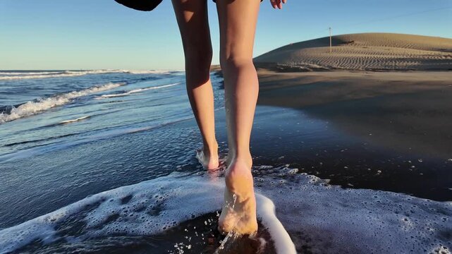 Woman walking barefoot on a sandy beach at sunset, her feet splashing in the shallow ocean water as she strolls along the shoreline holding her sandals in her hand