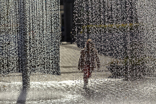 Europe heatwave concept with blurred child running through city fountain. Summer weather crisis, extreme heat wave climate change impact. Water droplets splashing, urban refreshing. Global warming